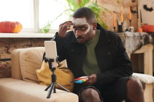 Influencer marketing strategy of a serious young Afro-American man in black jacket sitting in armchair and shooting video on smartphone how to paint face for Halloween party